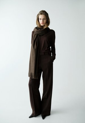 Femme debout devant un fond uni, portant un pantalon large marron foncé, un haut assorti, des chaussures noires pointues et une longue écharpe marron.