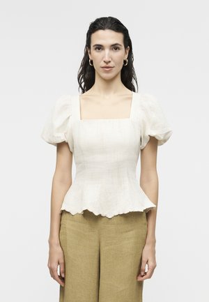 Γυναίκα με σκούρα μαλλιά, φορούνταν λευκό πουκάμισο με φουσκωτά μανίκια και μπεζ παντελόνι ψηλής μέσης, στέκεται μπροστά σε ένα απλό ανοιχτόχρωμο φόντο.