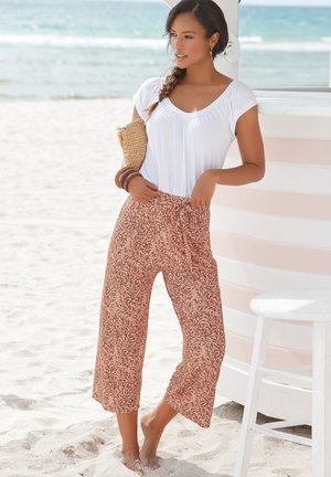 White short-sleeve top and patterned, wide-leg pants in rust and cream with a floral design. Worn with stacking bracelets, standing on sand.