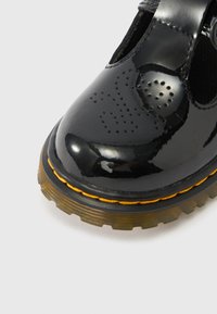 Chaussure en cuir verni noir avec détails perforés, bout arrondi et coutures jaunes. Semelle épaisse et texturée pour plus de profondeur et d'adhérence.
