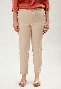 Pantaloni beige realizzati con un tessuto morbido ed elastico, con una vestibilità affusolata e una cerniera in vita; il modello è abbinato a sandali dorati.