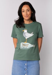 Groene katoenen t-shirt met een grafisch ontwerp van een meeuw in een gestreept shirt die op een fles staat met een papieren bootje erin.