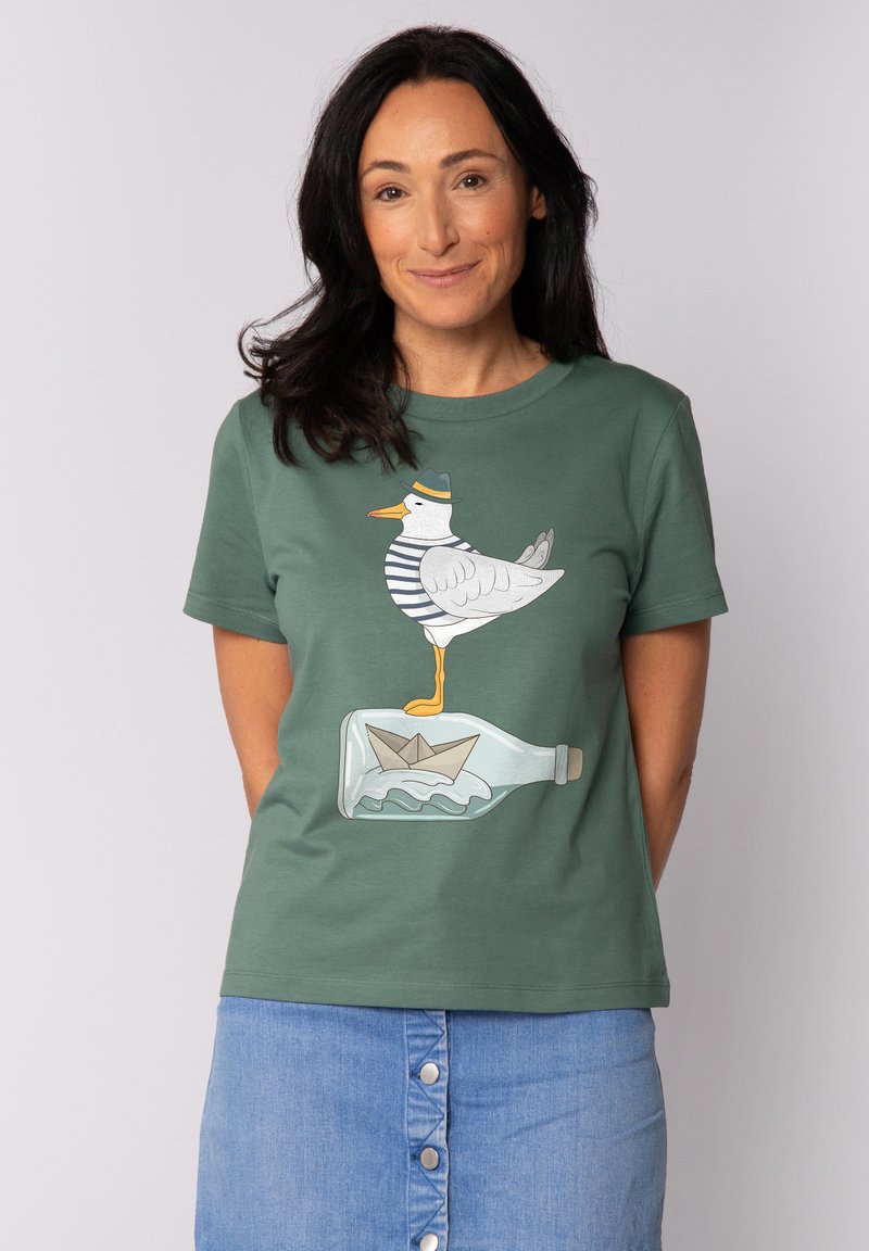 Groene katoenen t-shirt met een grafisch ontwerp van een meeuw in een gestreept shirt die op een fles staat met een papieren bootje erin.