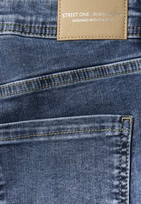 Jeans de mezclilla con un lavado azul claro, que presentan costuras amarillas, una etiqueta de cuero texturizada etiquetada como "Street One - Denim Atelier" y detalles en los bolsillos.