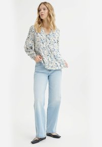 Helle, floral Bluse mit langen Ärmeln, V-Ausschnitt und gefaltetem Design, kombiniert mit hellblauen geraden Jeans und schwarzen Ballerinas.
