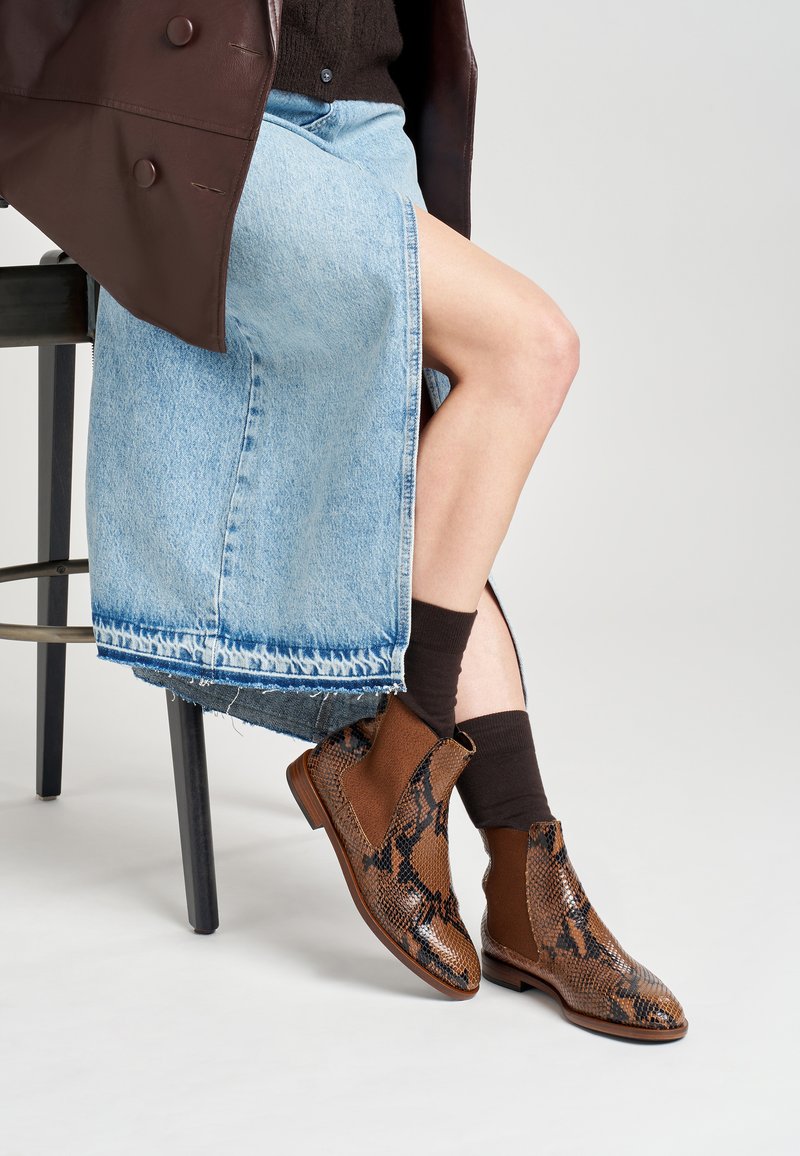 Botas de tobillo marrones con estampado de serpiente sintético y acentos de ante. Llevadas con una falda de mezclilla clara y calcetines oscuros, sentada en un taburete de metal.