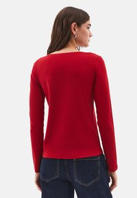 Kvinde med langt brunt hår iført sølv hoop øreringe, en rød bluse med lange ærmer og mørkeblåt jeans, vendt væk foran en hvid baggrund.