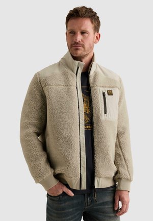 Homme portant une veste en polaire sherpa beige avec poche poitrine zippée, t-shirt noir à motif graphique et jean foncé, debout devant un fond uni.