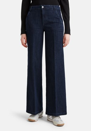 Donkerblauwe jeans met wijde pijpen, gedragen met witte sneakers en een zwart top met lange mouwen, getoond van taille tot voeten op een witte achtergrond.