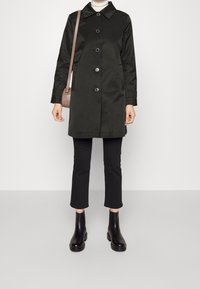 Person som bär en svart knäppt kappa, kortsvarta byxor, svarta ankelboots, vit polotröja och bär en brun axelväska.