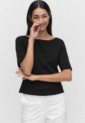 Haut noir à manches courtes avec un large col et une coupe droite, présentant une texture douce. Associé à un pantalon de couleur claire.