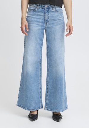 Persona che indossa jeans azzurri a gamba larga e scarpe nere con punta, in piedi contro uno sfondo semplice.