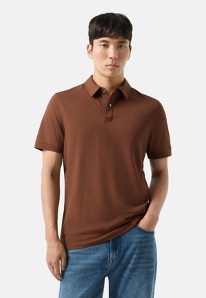 Ung mand iført en brun kortærmet polotrøje og blå jeans, stående med den ene hånd i lommen mod en ensfarvet baggrund.
