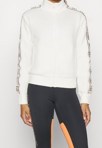 Chaqueta blanca con cremallera y cuello alto, con cinta de logo negra a lo largo de las mangas. Tejido suave, puños y dobladillo acanalados, diseño minimalista.