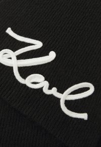 Schwarze gestrickte Mütze mit weißem gesticktem Text. Der Stoff wirkt weich mit einer gerippten Textur und verfügt über kunstvolle Schriftverzierungen.