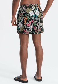 Homme portant un short de bain noir à motifs floraux et des sandales slides noires, debout avec une main sur la hanche sur un fond blanc.