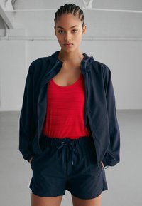 Giacca a vento navy con cappuccio, canotta rossa a coste e pantaloni corti navy con cordino. Tessuto morbido, vestibilità comoda, design funzionale.