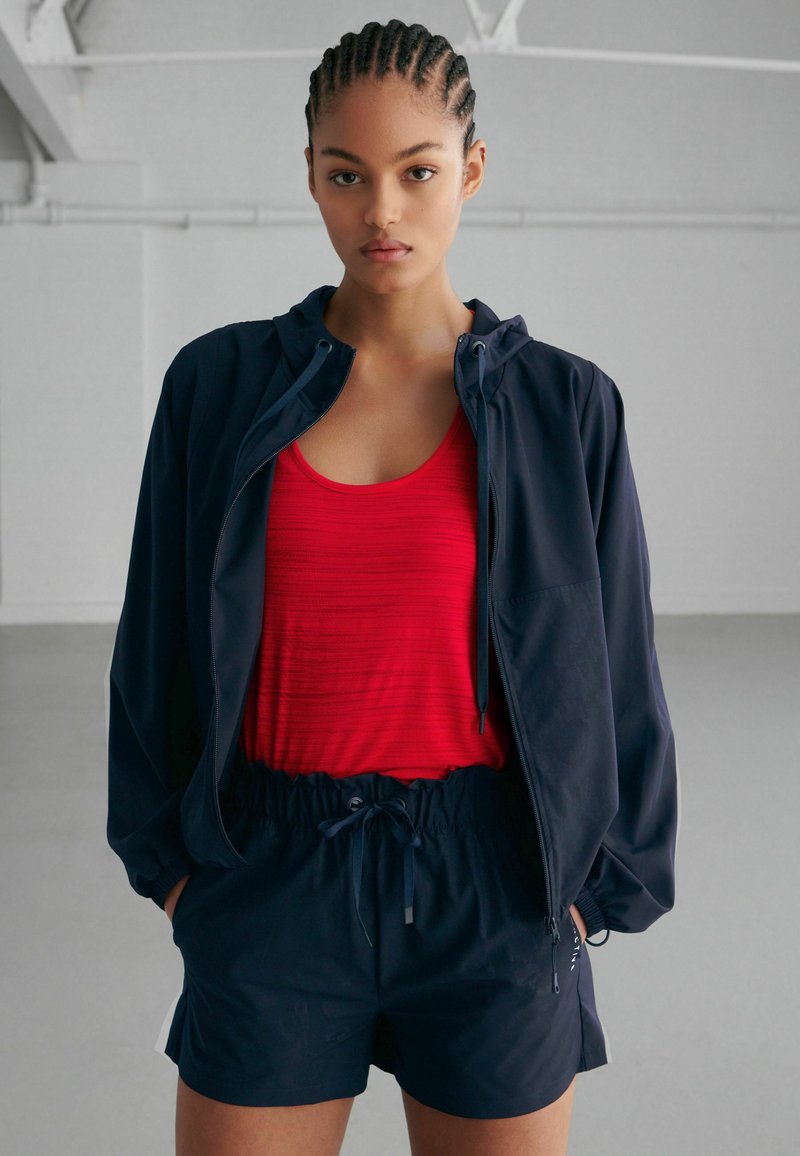 Giacca a vento navy con cappuccio, canotta rossa a coste e pantaloni corti navy con cordino. Tessuto morbido, vestibilità comoda, design funzionale.