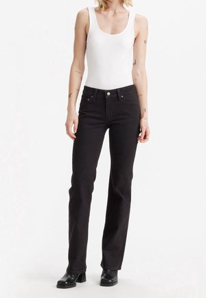 Vrouw draagt een witte mouwloze top, zwarte jeans met rechte pijpen en zwarte laarzen met hakken, staand tegen een eenvoudige witte achtergrond.