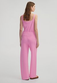 Roze gebreid overall met een ronde achterkant, wijde benen en een gestructureerde afwerking. Model staat in profiel tegen een neutrale achtergrond.
