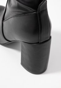 Botín de cuero negro hasta el tobillo con un tacón cuadrado y grueso. Presenta una textura lisa y detalles de costura a lo largo del caño para un diseño adicional.