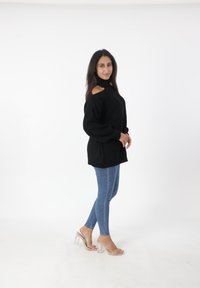Donna in piedi di lato che indossa un maglione nero con spalle a taglio, jeans blu e sandali con tacco alto a fascia trasparente su sfondo bianco.