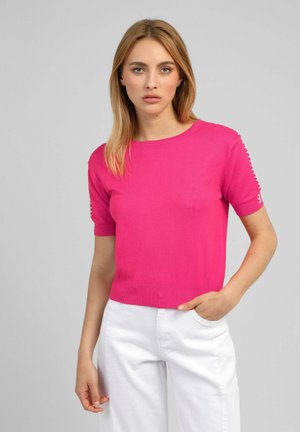 Femme aux cheveux blonds portant un haut rose vif à manches courtes et un pantalon blanc, debout devant un fond gris uni.