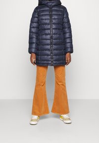 Marinblå pufferjacka med dragkedja i fronten, som har huva och horisontellt quiltat mönster, kombinerad med orange kordflossbyxor med utsvängd ben och vita sneakers.