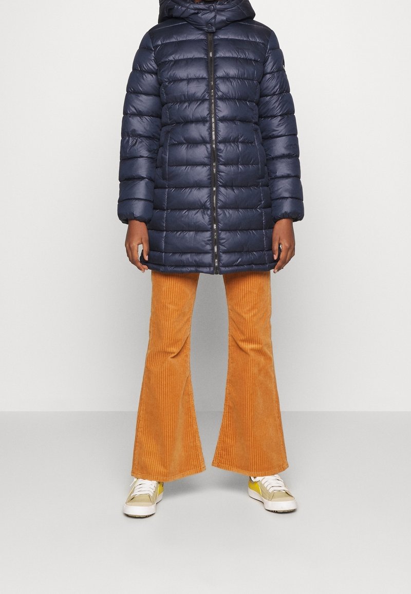 Marinblå pufferjacka med dragkedja i fronten, som har huva och horisontellt quiltat mönster, kombinerad med orange kordflossbyxor med utsvängd ben och vita sneakers.