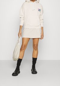 Béžová mikina s klokankovou kapsou a logem, doplněná o sladěnou sukni. Černé ponožky a robustní černé boty spolu s lehkou taškou přes rameno.