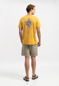 T-shirt di cotone gialla con una stampa di un orologio grigio sulla schiena, abbinata a pantaloni corti grigio chiaro e sandali marroni.