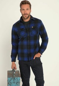 Geruit shirt in zwart en blauw, gemaakt van fleece. Heeft een knoopsluiting aan de voorkant, lange mouwen en een borstzak. Draagt het met een donkerdenim en houdt een metalen doos vast.