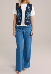 Laikrasčių siuvinėtas jūrinės spalvos liemenė, derinama su balta marškinėliais ir šviesiai mėlynomis plačiomis kelnėmis. Sandalai užbaigia atsitiktinį ansamblį.