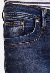 Jeansy z ciemnoniebieskiego denimu o postrzępionej fakturze, z widocznymi przeszyciami i przednią kieszenią. Elementy ozdobne to metalowy guzik i ćwieki.