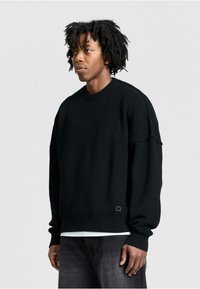 Black knit sweater with a round neckline and loose fit. Features ribbed cuffs and hem, textured pattern, and subtle logo detail at the bottom.