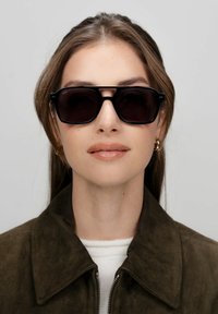 Zwarte rechthoekige zonnebril met donkere lenzen, voorzien van een stevig montuur en een minimalistisch ontwerp. Gekleed door een model in een groene leren jas.