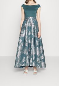 Robe teal avec décolleté épaules nues ; la jupe présente un motif abstrait argent métallique ; le mannequin porte des sandales bleu clair.