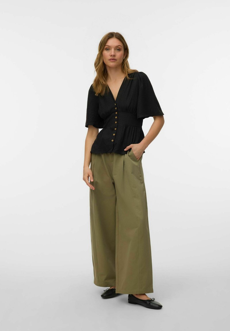 Kobieta stojąca, ubrana w czarną koszulę na guziki z dekoltem w serek, szerokimi rękawami oraz luźne spodnie o szerokich nogawkach w kolorze oliwkowej zieleni, do tego czarne baleriny.