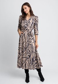 Długa, zwężająca się sukienka w kolorze beżowym z czarnym wzorem zebry. Dekolt w serek, bufiaste rękawy, boczne kieszenie i marszczona talia. Noszona z czarnymi kozakami za kolano.