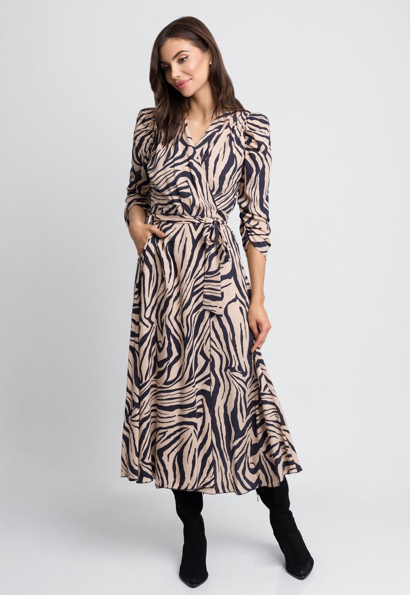 Długa, zwężająca się sukienka w kolorze beżowym z czarnym wzorem zebry. Dekolt w serek, bufiaste rękawy, boczne kieszenie i marszczona talia. Noszona z czarnymi kozakami za kolano.