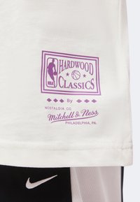 Λευκό ύφασμα με μοβ λογότυπο NBA "Hardwood Classics" και "Mitchell & Ness Philadelphia, PA" κείμενο πάνω από μαύρο σορτς με λευκή «φτερούγα» Nike.
