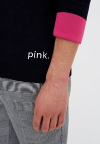 Braccio di una persona in un maglione nero con polsini rosa e il testo "pink." bianco, appoggiato accanto a pantaloni a scacchi bianchi e neri.