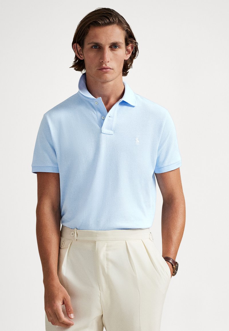 Svetlomodré polo tričko s textúrovanou látkou, krátkymi rukávmi a dvoma gombíkmi na zapínanie. Na hrudi je malé biele logo.