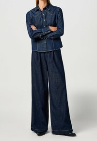 Henkilö, joka on pukeutunut tummansiniseen farkkupaitaan pitkissä hihoissa, jossa on helmipainikkeet, ja leveisiin farkkuihin, seisoo käsivarret ristissä.