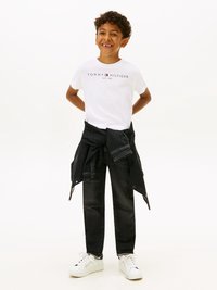 Λευκό βαμβακερό μπλουζάκι με λογότυπο "Tommy Hilfiger", συνδυασμένο με μαύρα τζιν και ένα σκούρο τζιν τζάκετ δεμένο γύρω από τη μέση. Λευκά αθλητικά παπούτσια.