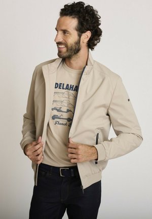 Homme aux cheveux bouclés sourit en regardant vers la gauche, portant une veste beige sur un t-shirt graphique beige et un jean foncé sur un fond uni.