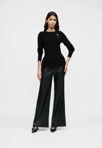 Pull en maille noire avec une taille à basque, assorti à un pantalon en cuir noir à jambes larges. Le pull est orné d'une petite broche florale bleue.