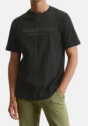 Mann trägt ein schwarzes Marc O'Polo T-Shirt mit dem Schriftzug „EST. IN STOCKHOLM“ und olivgrüne Hose, eine Hand in der Tasche, vor weißem Hintergrund.