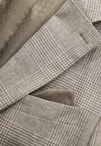 Detailný záber na vrecko béžovo-hnedého károvaného saka s vreckovkou so zladeným vzorom vo vnútri.