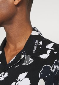 Camisa negra con estampado floral blanco, cuello corto y marca "CALVIN KLEIN" en el cuello. La tela parece suave y ligera.